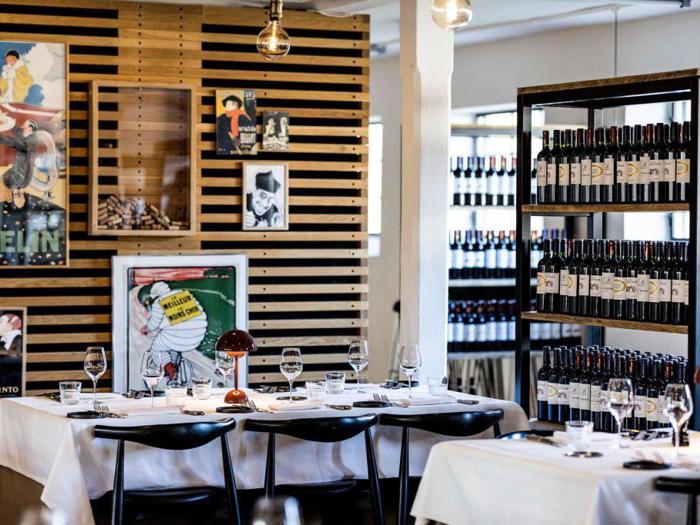 En moderne restaurant med hvide duge, sorte stole, vinflasker på hylder og kunstplakater på en væg af trælameller. Bordene er dækket med glas, tallerkener og servietter.