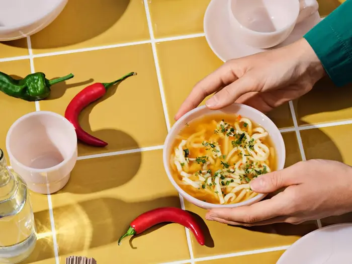 Hænderne holder en skål nudelsuppe på et gult flisebord med røde og grønne chilipeber, kopper, tallerkener og en vandflaske.