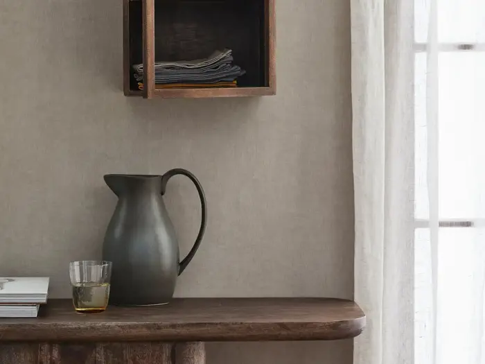 En mørk keramikkande, et glas med en lys drink og bøger står på et træbord under en væghylde med foldet sengetøj ved siden af et gennemsigtigt gardin, som sollyset skinner igennem.