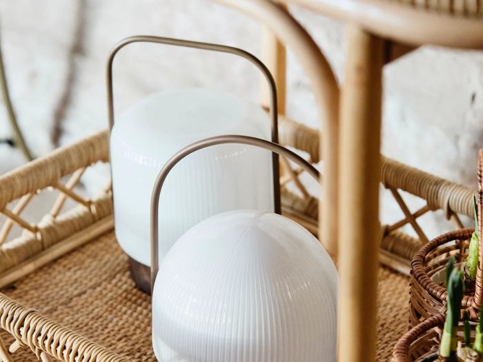 To moderne hvide lanterner med buede metalhåndtag står på en flettet rattanbakke, og en del af en flettet kurv og møbler er synlige i nærheden.