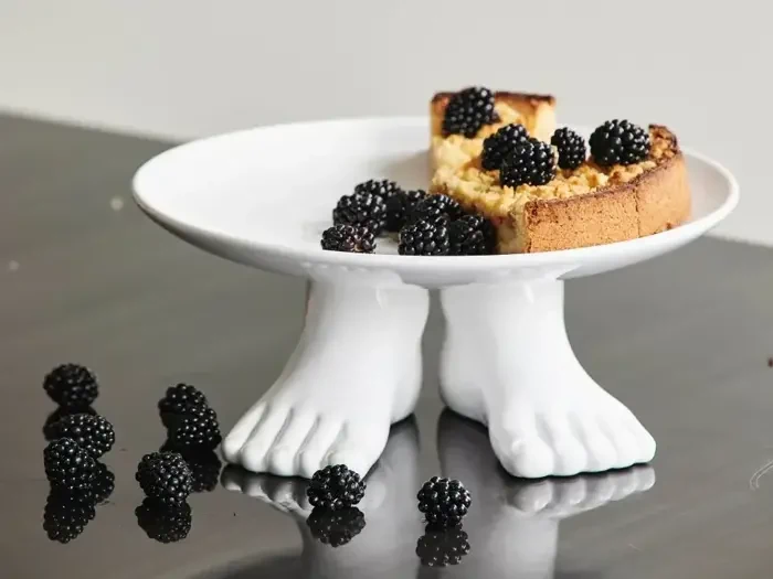 En kage med brombær på en hvid kageopsats formet som to fødder, med flere brombær spredt på en mørk overflade.