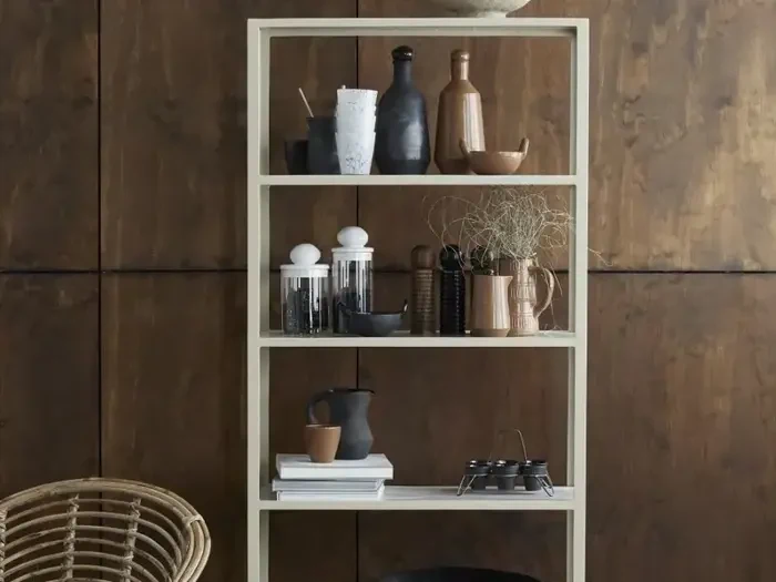 På en beige reol står forskelligt keramik, krus, vaser og skåle pænt opstillet mod en brun trævæg. En kurvestol er delvist synlig ved siden af.