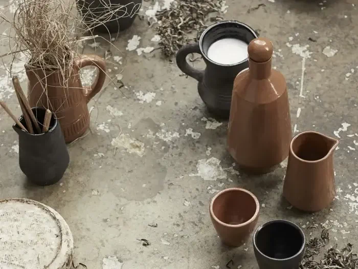 Forskellige keramiske kopper, kander og en vase med tørrede kviste arrangeret på en forvitret betonoverflade. Nogle kopper indeholder væske, andre indeholder pensler eller er tomme.