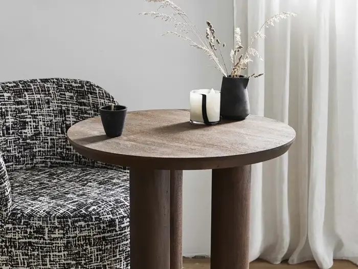 Et rundt træbord med en sort vase med tørret græs, et hvidt stearinlys og en sort kop ved siden af en mønstret sort-hvid stol. Hvide gardiner hænger i baggrunden.