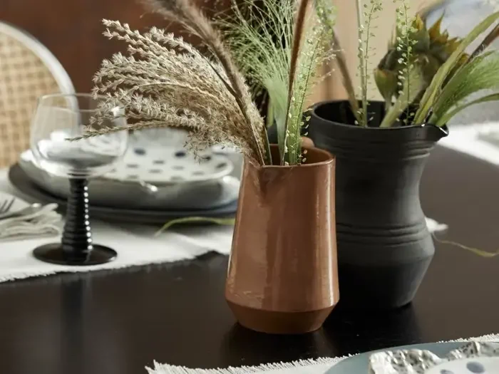 En brun keramikvase med tørret græs og en sort kande med grønt står på et mørkt bord dækket med tallerkener, glas og servietter.