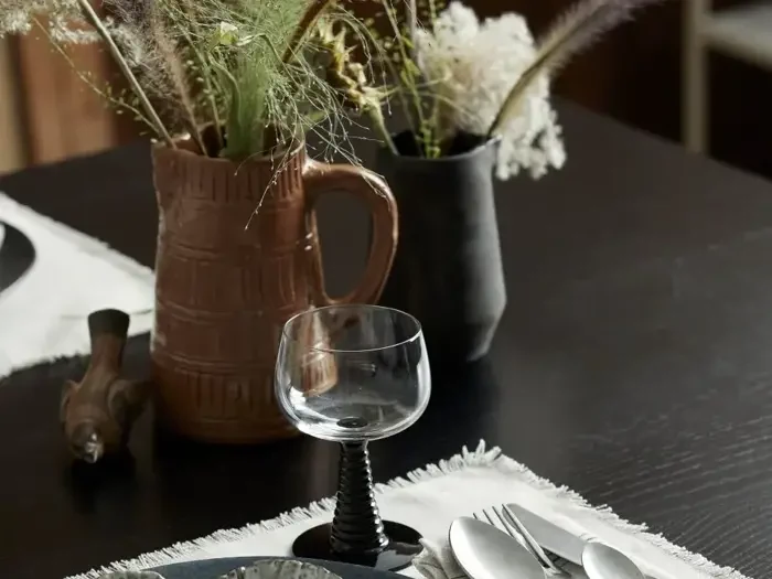 Nærbillede af et spisebord dækket med vinglas, bestik og tallerkener med keramikvaser med tørrede blomster i baggrunden.