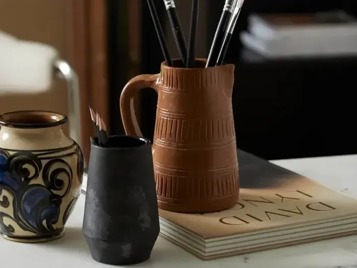 Tre keramiske kopper, den ene med sorte pensler, står på et bord ved siden af en lukket bog med titlen DAVID LYNCH med en sløret stol i baggrunden.