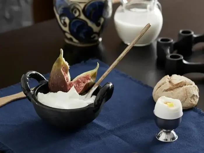 En sort skål yoghurt med frisk figen og ske står på et blåt klæde ved siden af et blødkogt æg, et rundstykke og en kande mælk.