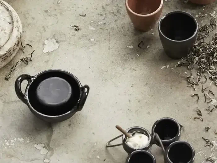 En række tomme keramiske kopper og skåle er anbragt på en ru, grå overflade ved siden af spredte, tørrede blade.