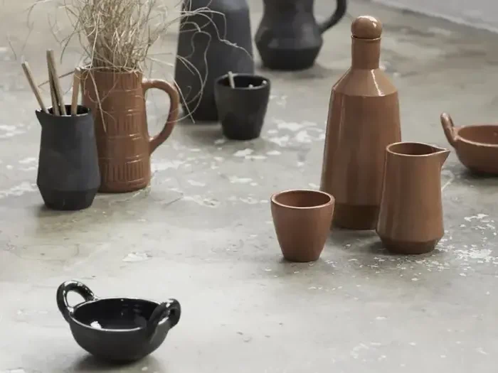 Forskellige keramiske vaser, kopper og kander i brune og sorte farver arrangeret på et struktureret betongulv. Nogle kar indeholder tørrede planter eller pinde.