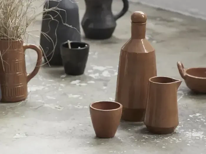 Brune keramikkopper, en kande og en flaske står på et betongulv med andet keramik og en kande med tørret græs i baggrunden.