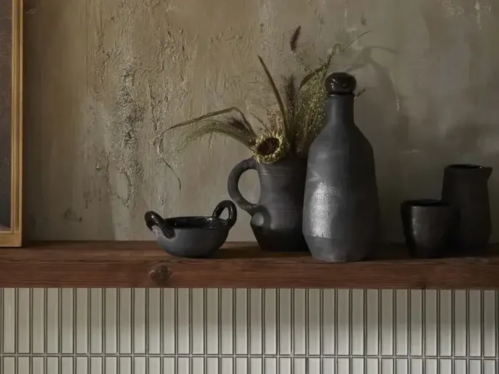 På en træhylde står mørke keramikkar og en kande med tørret græs mod en struktureret beige væg og et hvidt flisebelagt backsplash.