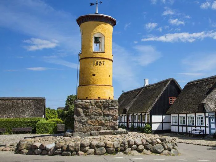 Et gult, cylindrisk tårn med årstallet 1857 står på en stenbase, omgivet af traditionelle stråtækte huse under en blå himmel.