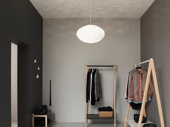 Minimalistisk gang med en moderne hvid pendellampe, tøjstativer i træ med hængende tøj og en mørk accentvæg med små pyntegenstande.