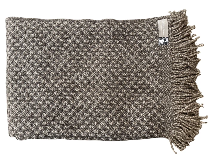 Et foldet, struktureret gråt og hvidt strikket tæppe med frynsede kanter og et mærke i øverste højre hjørne.