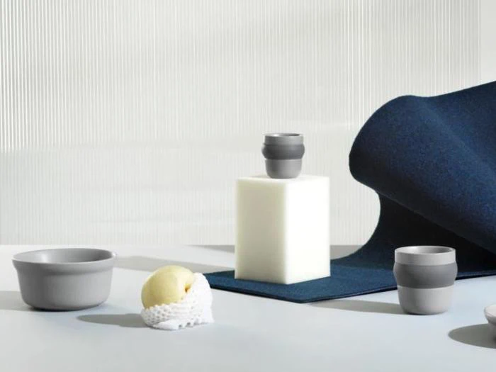 Minimalistisk scene med grå keramiske kopper og skåle, en kugle is i et hvidt net, en hvid rektangulær blok og mørkeblåt stof på en lys baggrund.