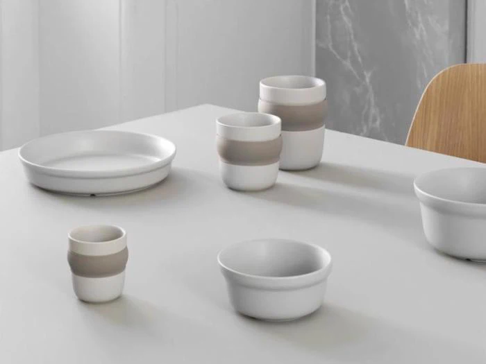 En minimalistisk borddækning med hvide keramiske fade og kopper, nogle med beige bånd, arrangeret på et lyst bord i en moderne indretning.