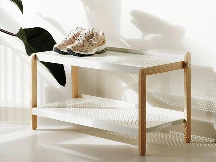 En minimalistisk skoreol i hvid og træ med to hylder, hvor der står et par beige sneakers på den øverste hylde. Sollys og planteskygger er synlige i baggrunden.