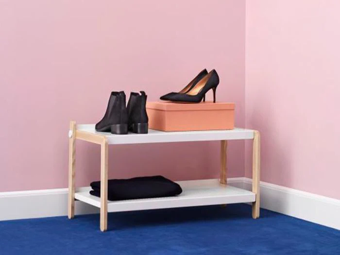 Et lille skostativ i to etager rummer et par sorte hæle på en kasse og ankelstøvler på toppen, med sammenfoldet tøj på den nederste hylde, opstillet mod lyserøde vægge og et blåt tæppe.