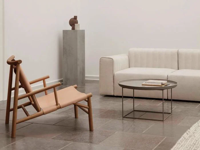 Minimalistisk stue med en beige sofa, en træstol, et rundt sofabord med en bog, klinkegulv og en høj grå piedestal med en lille abstrakt skulptur.
