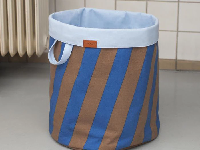 En vasketøjskurv af stof med blå og brune lodrette striber, en lyseblå kant og et lille håndtag, placeret på et gråt gulv i nærheden af en radiator og en flisebelagt væg.