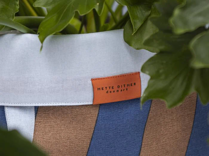 Et nærbillede af en stofkrukke med blå og brune striber, hvid kant og en brun etiket med teksten METTE DITMER denmark, omgivet af grønne blade.