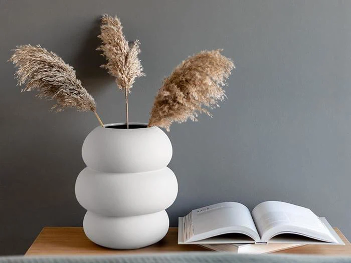 En moderne hvid vase med tre stængler af pampasgræs står på en træplade ved siden af en åben bog på en grå væg.