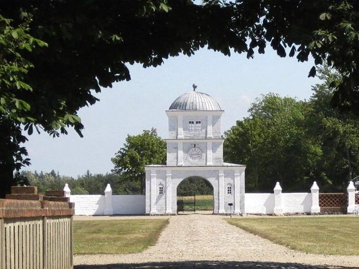 Et hvidt porthus med ur og kuppeltag står for enden af en træbelagt sti, indrammet af grønne grene og omgivet af en hvid mur.