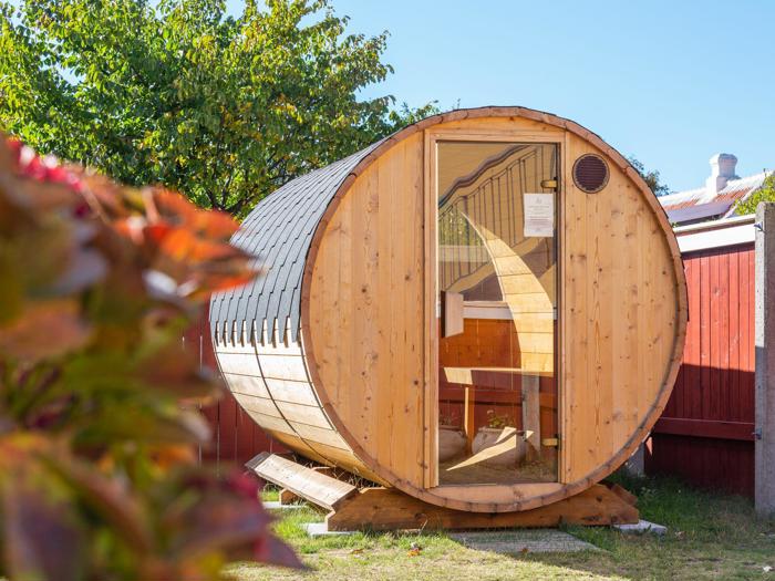 En tøndeformet sauna af træ står udendørs på græs med en glasdør, der viser bænke indeni. I baggrunden ses træer og et rødt hegn.