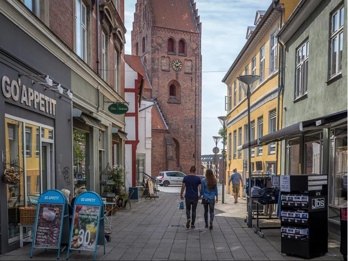 Et par går ned ad en gågade med butikker og udendørs udstillinger med en høj murstenskirke og et klokketårn i baggrunden.