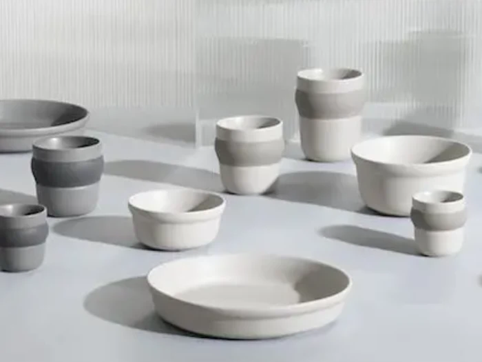 En samling af moderne, minimalistiske keramiske skåle og kopper i hvide og grå nuancer, arrangeret på en lys overflade med en blød baggrund.