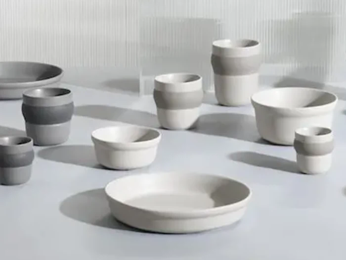 Forskellige keramiske skåle og kopper i hvide og grå toner er arrangeret på en flad overflade med blød belysning og skygger.