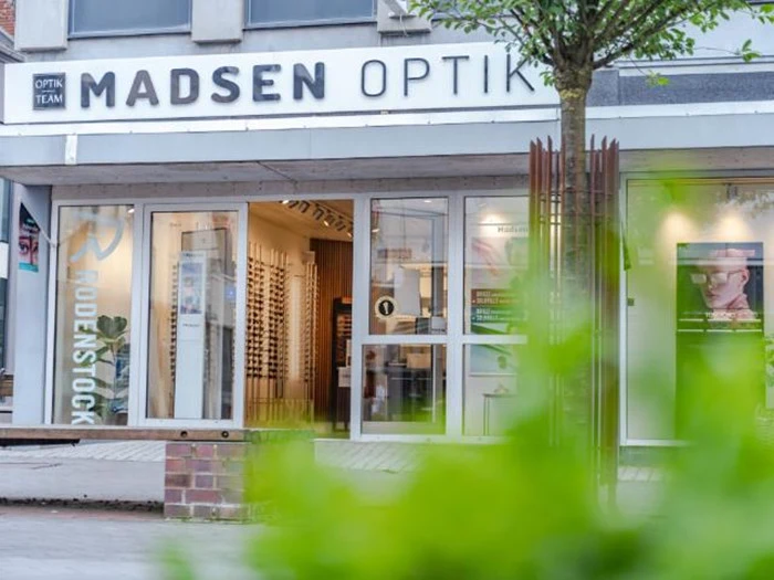 Gadebillede af Madsen Optik, en brillebutik med store glasvinduer, udstillingshylder med briller og en grøn busk i forgrunden.