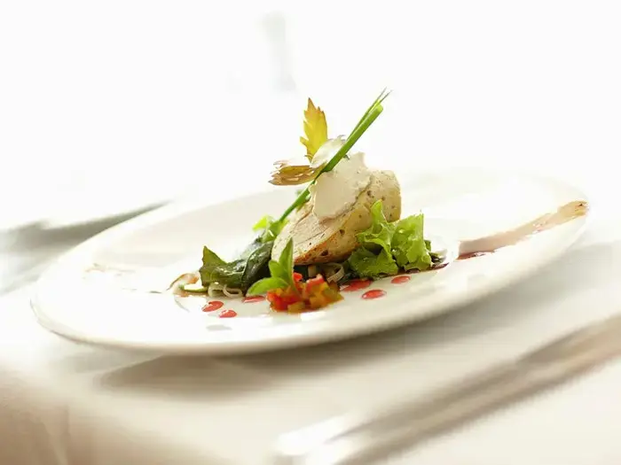 En gourmetret med et stykke fisk på blandet grønt, pyntet med krydderurter og sauce, serveret på en hvid tallerken med kunstnerisk præsentation.