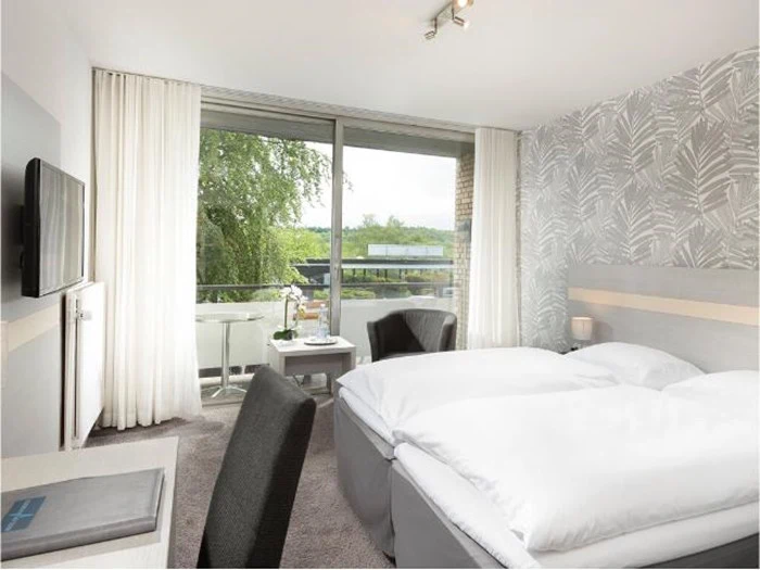 Moderne hotelværelse med to enkeltsenge, mønstret accentvæg, skrivebord, stole, tv og stort vindue med gennemsigtige gardiner med udsigt til en balkon og grønne områder udenfor.