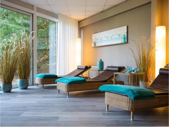En moderne spa-lounge med flettede chaiselonger, blågrønne håndklæder, høje potteplanter, store vinduer og dæmpet belysning.
