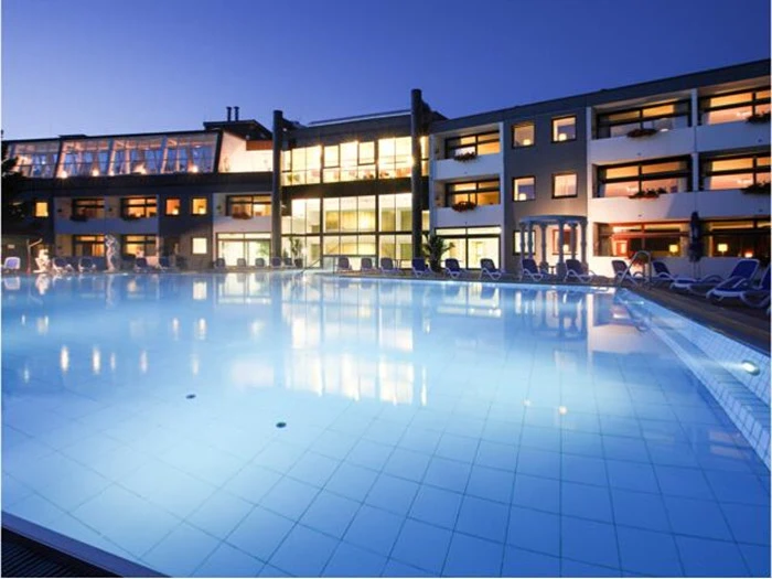 En stor, oplyst udendørs swimmingpool foran en moderne hotelbygning i flere etager i skumringen.