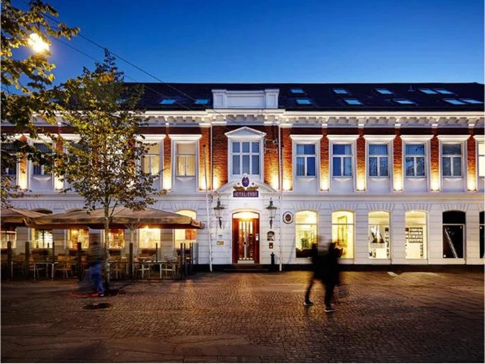 Et veloplyst hotel i to etager med udeservering, store vinduer og mennesker, der går forbi på en brostensbelagt gade i skumringen.