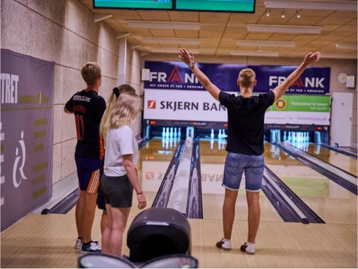Fire unge mennesker står ved en bowlingbane; den ene med armene i vejret for at fejre, mens de andre holder øje med banerne.