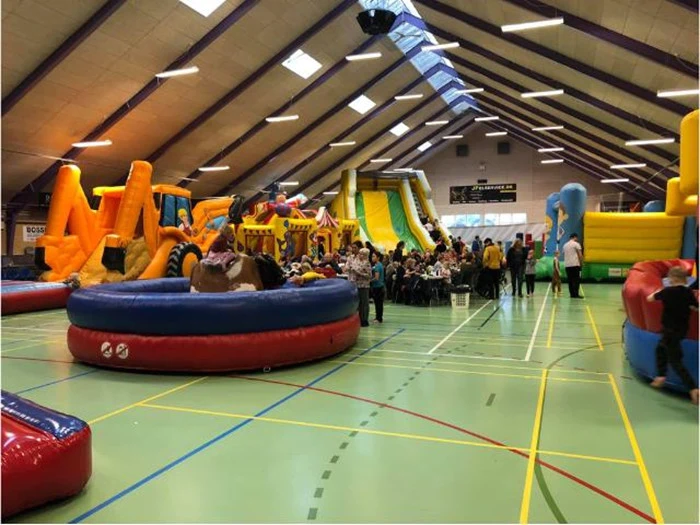 En stor indendørs gymnastiksal fyldt med farverige oppustelige rutsjebaner og hoppeborge; børn og voksne leger og går rundt.