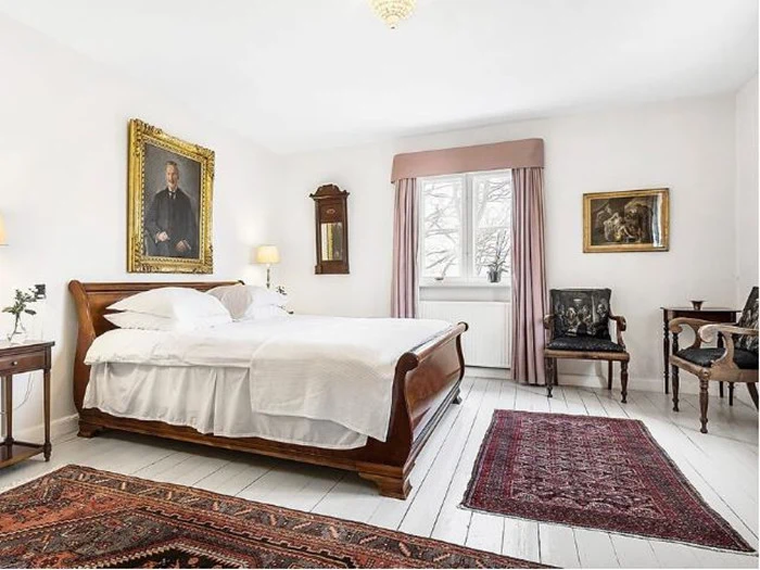 Et lyst, klassisk soveværelse med en køjeseng af træ, hvidt sengetøj, antikke møbler, indrammet kunst og mønstrede tæpper på hvide trægulve. Sollyset strømmer ind gennem et vindue med gardiner.