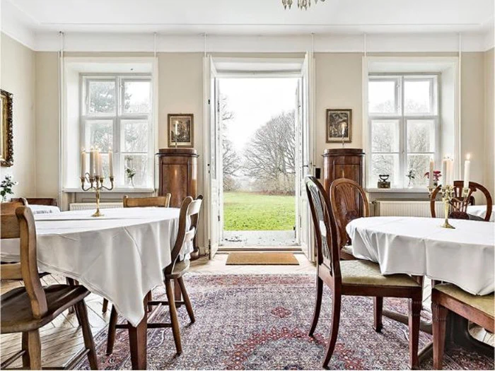 En lys spisestue med runde borde dækket af hvide duge, træstole og store vinduer med en dør ud til en græsklædt gård og træer udenfor.