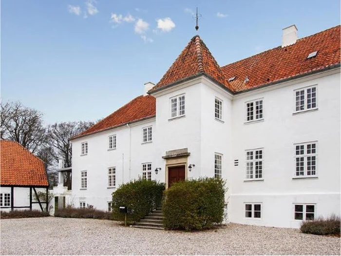 Et stort hvidt hus i to etager med rødt tegltag, flere vinduer og en central indgang med trappe, omgivet af grus og buske.