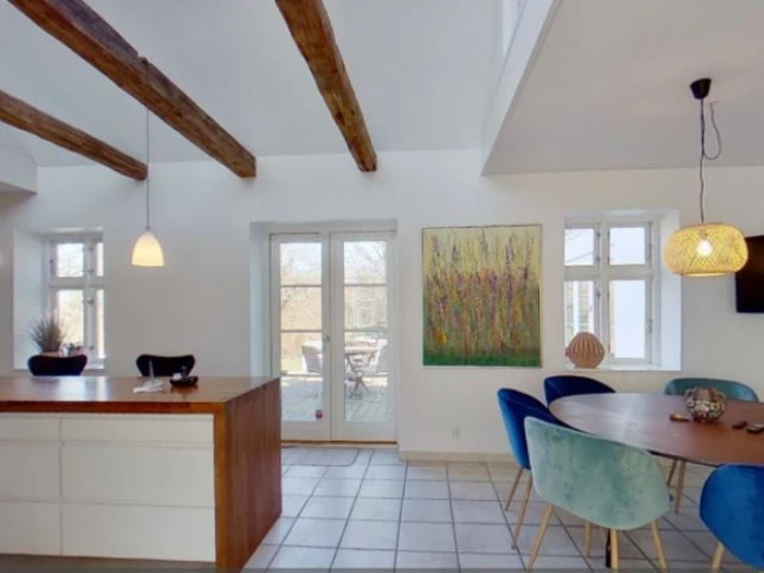 En lys spiseplads med synlige træbjælker, et rundt bord med blå stole, pendellamper og et stort maleri på væggen ved siden af glasdøre, der fører udenfor.