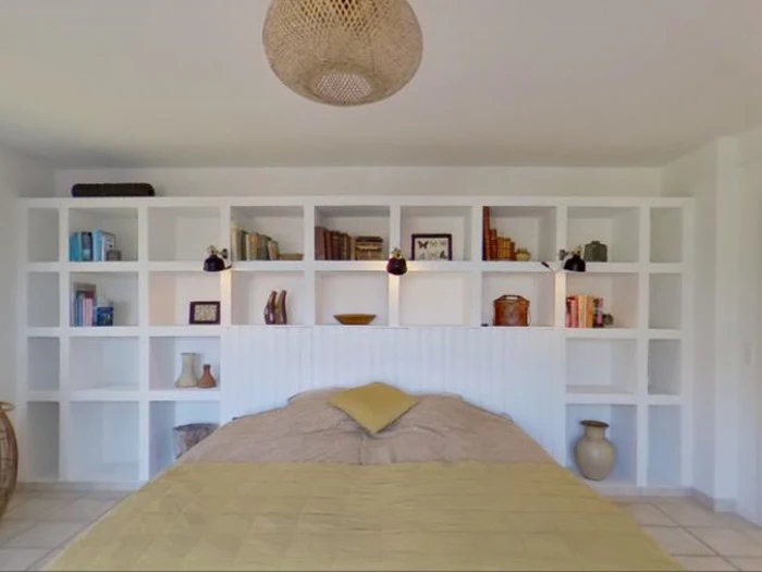 Et hyggeligt soveværelse med et beigefarvet sengetæppe, en gul pude og en stor hvid indbygget bogreol fyldt med bøger og dekorationer bag sengen. En vævet loftslampe hænger over hovedet.