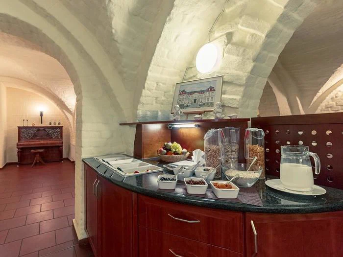 Morgenmadsbuffet med korndispensere, skåle med frugt, nødder og mælk på en sort disk i et rum med buede hvide murstenslofter og klinkegulv.