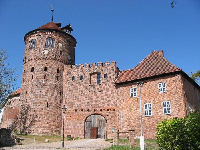 Et historisk murstensslot med et stort rundt tårn, buede trædøre og røde tegltage mod en klar blå himmel.