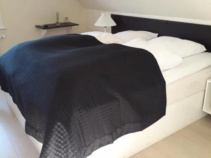 En pænt redt seng med fire hvide puder og et sort quiltet tæppe i et minimalistisk soveværelse med lyse vægge og en lille hylde med en plante og et glas.