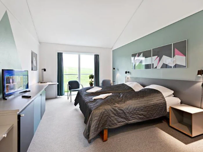 Moderne hotelværelse med to senge, gråt sengetøj, tv, skrivebord, kunstværker over sengene, stort vindue med gardiner, to stole og et lille bord ved vinduet.