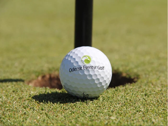 En golfbold med Odense Eventyr Golfs logo hviler på kanten af et hul på en pænt trimmet green.